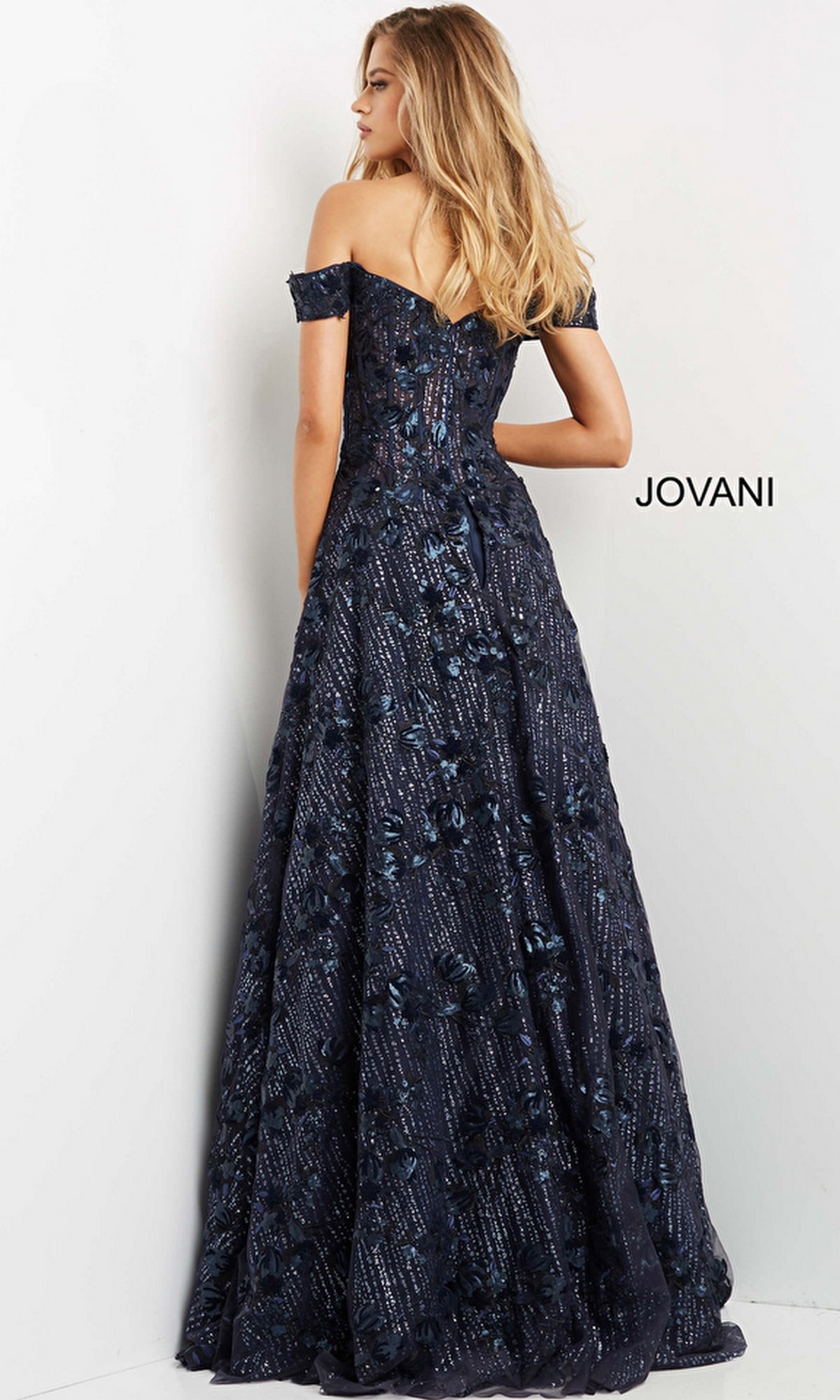 A woman wearing the Long Prom Dress 07162 by Jovani—navy blue, off-the-shoulder, with a floral pattern—stands facing a white wall displaying the word